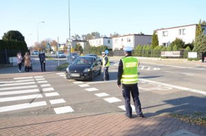 Policjant stoi przed przejazdem rowerowym , natomiast drugi policjant stoi na jezdni, rozmawia z kierowcą czarnego samochodu osobowego, który chce wjechać w boczną ulicę.