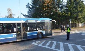 Policjant stoi na jezdni, tyłem do samochodów, które zatrzymały się na drodze za nim, przed nim przez skrzyżowanie przejeżdża radiowóz.