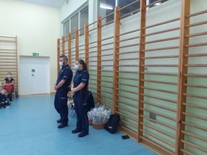 Na sali gimnastycznej dwoje policjantów prowadzi spotkanie z dziećmi, widać w tle policjantów drabinki ścienne a po prawej stronie policjantów widoczne są dwie osoby, uczeń i osoba dorosła.