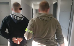 Policjant prowadzi zatrzymanego przez korytarz, zatrzymany mężczyzna na rękach ma założone kajdanki.