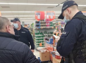 Policjant rozmawia z mężczyzną w sklepie. Ma to związek z tym, że nie miał on maseczki. Twarz mężczyzny jest zapikslowana.