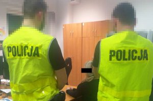 Dwóch policjantów stoi a za nimi siedzi zatrzymana kobieta.