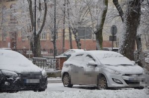 Na parkingu stoją zaparkowanie koło siebie dwa samochody osobowe, są pokryte śniegiem na dachu, masce i szybach.