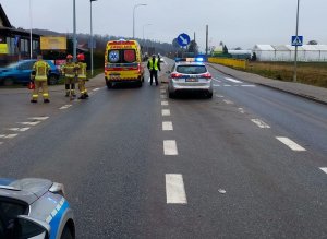 Radiowóz stoi na pasie ruchu, obok niego karetka, pomiędzy stoi policjant. Na poboczu po lewej stronie stoi przy ogrodzeniu niebieski rozbity samochód.