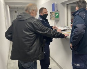 Policjanci stoją z mężczyzną przy okienku, przy którym jest lada i na niej dokumenty. Pierwszy z policjantów trzyma w ręku urządzenie, służące do pomiaru zawartości alkoholu. Mężczyzna ma zsuniętą maseczkę z ust, na potrzeby przeprowadzenia badania trzeźwości.