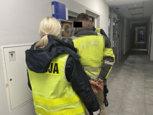 Policjanci stoją przy oknie i ladzie, między nimi stoi zatrzymany mężczyzna, który zostanie poddany badaniu trzeźwości.