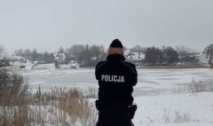 Policjant stoi nad brzegiem zamarzniętego jeziora. Wszystko w koło jest zaśnieżone a jezioro zamarznięte.