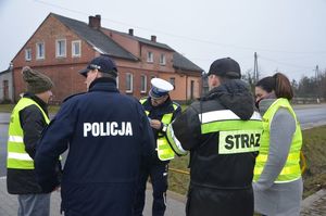 Na zdjęciu stoi grupa osób na prost policjant w granatowej kurtce z napisem POLICJA,obok niego po prawej stronie stoi strażak, a dalej koło strażaka stoi policjantka w kamizelce odblaskowej z napisem POLICJA, obok policjanta w granatowej kurtce służbowej po lewej tronie stoi przedstawiciel zarządcy drogi a na wprost stoi policjant ruchu drogowego.