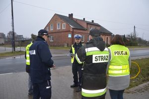 Na zdjęciu przy drodze stoi grupa osób na prost policjant w granatowej kurtce z napisem POLICJA,obok niego po prawej stronie stoi strażak, a dalej koło strażaka stoi policjantka w kamizelce odblaskowej z napisem POLICJA, obok policjanta w granatowej kurtce służbowej po lewej tronie stoi przedstawiciel zarządcy drogi a na wprost stoi policjant ruchu drogowego.