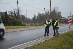 W pewnym oddaleniu na drodze stoi policjant ruchu drogowego, obok niego stoi przedstawiciel zarządcy drogi a dalej za nimi policjant w granatowym umundurowaniu i czapce z daszkiem.