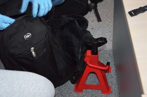 Na podłodze leży lewarek do samochodu obok dwa czarne plecaki, które są w trakcie rozpakowywania przez policjanta.