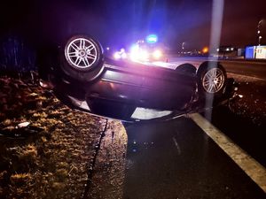 Samochód leży na poboczu drogi na dachu, w tle widoczne pojazdy z włączonymi sygnałami świetlnymi.