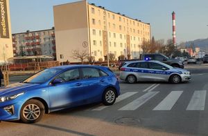 Samochód osobowy kia w kolorze niebieskim stoi częściowo na przejściu dla pieszych, za nim stoi policyjny radiowóz