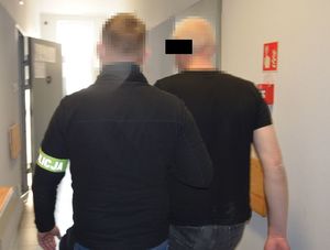 Zatrzymany mężczyzna jest prowadzony korytarzem. Policjant na ramieniu ma założoną opaskę z napisem POLICJA.