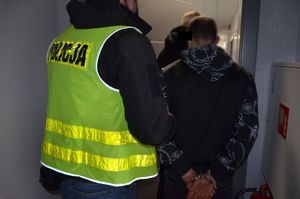 Policjant prowadzi zatrzymanego, który ma kajdanki założone na ręce trzymane z tyłu