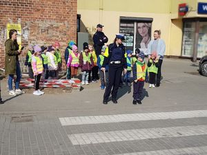 Policjantka stoi przed przejściem dla pieszych obok niej i za nią stoi dzielnicowy wraz z grupą przedszkolaków.