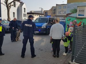 Policjant stoi przy budynku, w którym mieście się przedszkole, z jednej strony obok niego stoją przedszkolaki a na przeciwko policjant przewodnik wraz z psem patrolwo-tropiącym oraz w tle widoczny jest radiowóz.