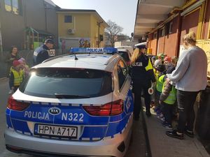 Na pierwszym planie stoi radiowóz obok niego stoją policjanci oraz dzieci, których część zagląda do środka.