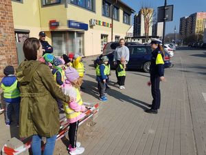 Policjantka stoi tyłem do przejścia dla pieszych, przodem do dzieci, którym objaśnia bezpieczne zachowanie podczas przechodzenia przez jezdnię.