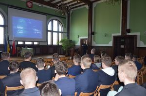 Policjant prowadzi spotkanie, przemawia do uczniów, w tle na ekranie multimedialnym wyświetlono zdjęcie fragmentu radiowozu z napisem Policja.