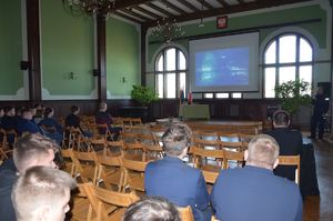 Policjant prowadzi spotkanie, przemawia do uczniów, w tle na ekranie multimedialnym wyświetlono zdjęcie fragmentu radiowozu z napisem Policja.