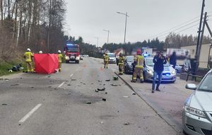 Technik fotografuje samochód, którego fragment jest po prawej stronie zdjęcia, a po lewej na poboczu widoczny jest motocykl oraz parawan czerwony z napisem STRAŻ. Na drodze stoją strażacy i policjant.