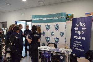 Policjanci rozmawiają z uczniami na holu szkolnym. W tle widoczny jest policyjny baner.