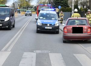 Po prawej stronie na drodze i częściowo na chodniku stoi samochód w kolorze czerwonym, a tuż za nim policyjny radiowóz. Na przodzie zdjęcia widoczne jest przejście dla pieszych.