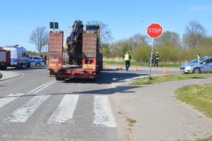 Na skrzyżowaniu stoi ciągnik rolniczy z naczepą - widoczny na zdjęciu jest tył naczepy, w pewnym oddaleniu po prawej stoi radiowóz. Widoczny jest znak stop oraz policjant stojący na drodze.