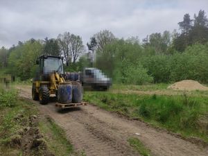 Ciągnik z podnośnikiem wiezie kilka beczek, w tle stoi samochód, na którym na przyczepce stoją beczki z chemikaliami.