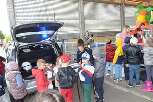 Policyjny radiowóz ma otwarty uniesiony do góry bagażnik, obok stoi stolik z policyjnymi plakatami a przy nim policjantka coś wypisuje na małych karteczkach, podchodzą do niej dzieci