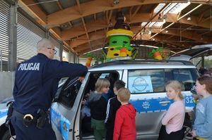 Policyjny radiowóz ma otwarte boczne drzwi, przy drzwiach przednich stoi policjant, który coś opowiada dzieciom wsiadającym do środka.