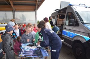 Policjantka stoi przy stoliku z policyjnymi plakatami i wypisuje coś na małych karteczkach, podchodzą do niej dzieci