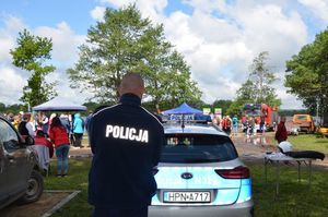 Policjant stoi na tle radiowozu a dalej za nim widoczni są zaproszeni goście na otwarcie przystani kajakowej.