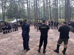 Policjantka stoi po lewej stronie, po jej prawej stronie stoi strażak a obok niego stoi mężczyzna, przed nimi i po lewej stronie stoją harcerze.