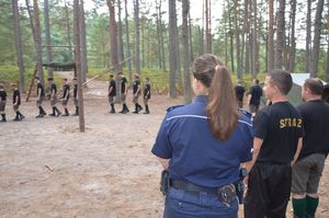 Policjantka stoi po lewej stronie, po jej prawej stronie stoi strażak a obok niego stoi mężczyzna, przed nimi i po lewej stronie stoją harcerze.