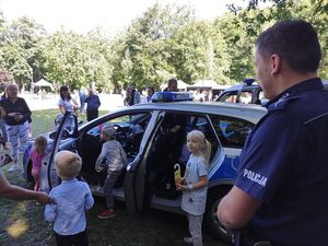 Policjant dzielnicowy stoi przy radiowozie i rozmawia z dziećmi i opiekunami.