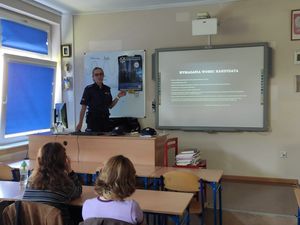 Policjantka stoi przy biurku, za nią widoczny jest plakat związany z doborem do służby w Policji, przed nią w ławkach siedzą uczniowie.