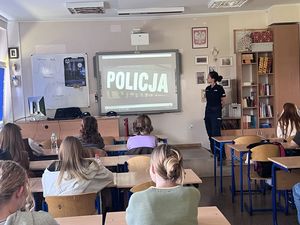 Policjantka przy tablicy multimedialnej, za nią na tablicy multimedialnej wyświetlony jest kadr z filmu o Policji, przed nią w ławkach siedzą uczniowie.