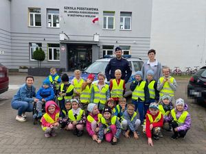 Policjant stoi razem z dziećmi przed szkołą, jest to zdjęcie grupowe, za nimi w tle widać także policyjny radiowóz.