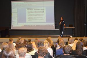 Policjantka prowadzi zajęcia, stoi na scenie w tle widać ekran, na którym wyświetla się prezentacja. Przed sceną siedzą uczniowie klas trzecich.