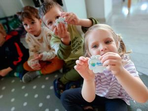 Dzieci siedzą na dywanie, w rękach trzymają - pokazują zawieszki odblaskowe.