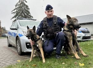 Policjant przykucnął z dwoma psami po bokach, po lewej stronie siedzi przy nim Luna, po prawej siedzi Boss.