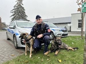 Policjant przykucnął z dwoma psami po bokach, po lewej stronie siedzi przy nim Luna, po prawej siedzi Boss.