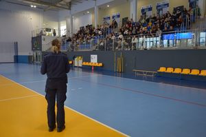 Policjantka prowadzi spotkanie profilaktyczne. Stoi na środku hali sportowej, młodzież siedzi  na trybunach w tle.