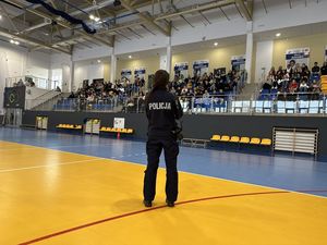 Policjantka prowadzi spotkanie profilaktyczne. Stoi na środku hali sportowej, młodzież siedzi  na trybunach w tle.