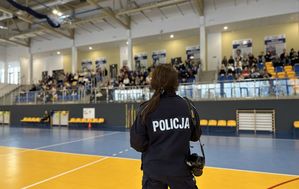 Policjantka prowadzi spotkanie profilaktyczne. Stoi na środku hali sportowej, młodzież siedzi  na trybunach w tle.