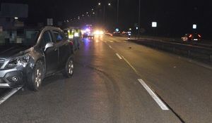 Na lewym poboczu przodem stoi samochód opel z rozbitym reflektorem. Za nim w tle poboczem idą dwaj policjanci w kamizelkach odblaskowych.