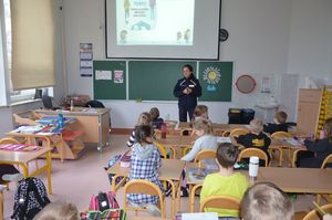 Policjantka prowadzi zajęcia, stoi przy tablicy, w tle wyświetlana jest prezentacja multimedialna.