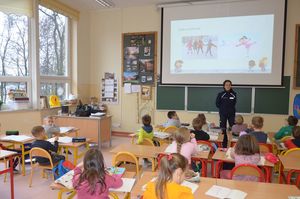 Policjantka prowadzi zajęcia, stoi przy tablicy, w tle wyświetlana jest prezentacja multimedialna.